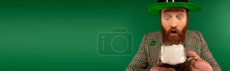 Shocked bearded man in hat looking at blurred glass of beer isolated on green, banner 