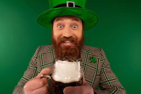 Fröhlicher bärtiger Mann mit Hut, Bier in der Hand und Blick in die Kamera während des Patronatsfestes isoliert auf Grün 