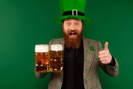 Excited man in hat with clover showing like and holding glasses of beer isolated on green 