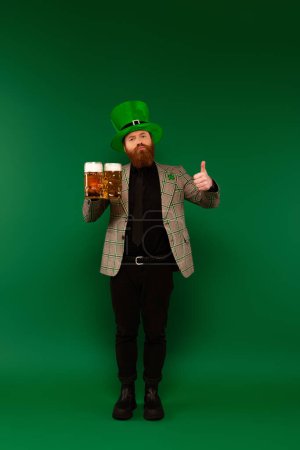 Bearded man in hat with clover holding glasses of beer and showing like on green background 