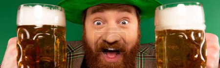 Excited bearded man holding glasses of beer and looking at camera isolated on green, banner 