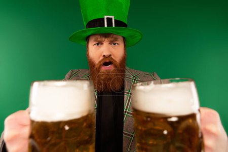 Schwerer bärtiger Mann mit Hut hält Biergläser isoliert auf Grün 