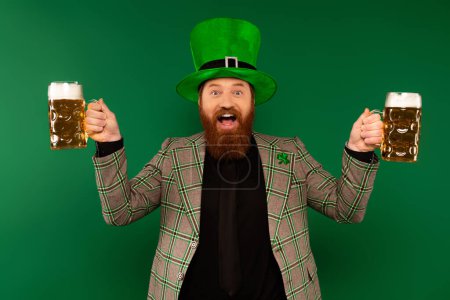 Positiver bärtiger Mann mit Hut feiert Patronatstag und hält Biergläser isoliert auf Grün 