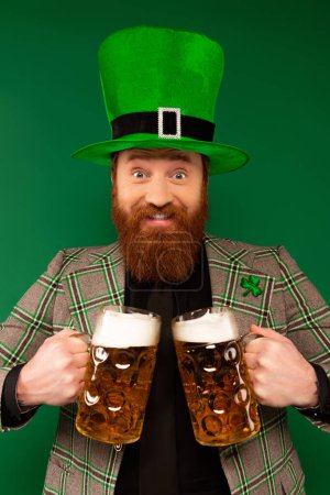 Lächelnder Mann mit Hut und Kleeblatt hält Biergläser isoliert auf Grün 