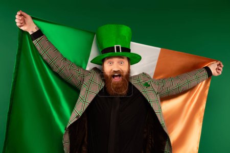 Excited bearded man in hat holding Irish flag isolated on green 