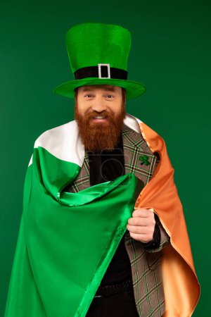 Hombre sonriente en sombrero con trébol envuelto en bandera irlandesa aislado en verde 