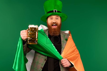 Un homme barbu joyeux tenant de la bière et un drapeau irlandais pendant la Saint Patrick isolé sur vert 