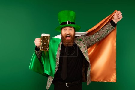 Hombre excitado en sombrero sosteniendo vaso de cerveza y bandera irlandesa aislado en verde 