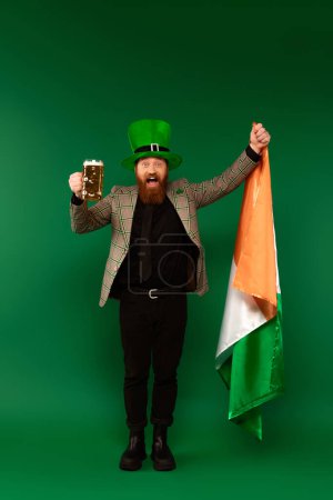 Hombre barbudo excitado con sombrero sosteniendo un vaso de cerveza y bandera irlandesa sobre fondo verde 