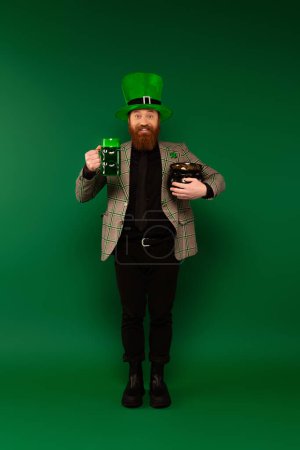 Happy bearded man in hat with clover holding glass of beer and pot with coins on green background 