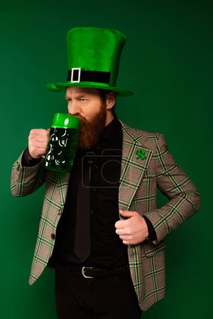 Serious man in hat with clover drinking beer during saint patrick day isolated on green 
