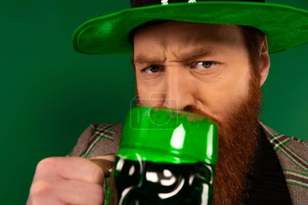 Foto de Retrato de un hombre serio en sombrero bebiendo cerveza aislado en verde - Imagen libre de derechos