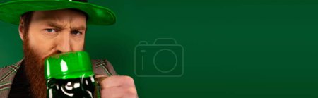 Téléchargez les photos : Serious man in hat drinking beer while celebrating saint patrick day isolated on green, banner - en image libre de droit