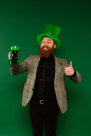 Smiling bearded man in hat holding beer and showing like isolated on green 