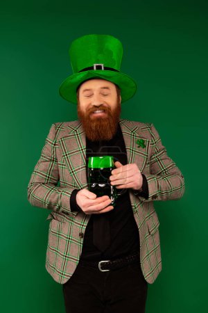 Joyful bearded man holding glass of beer while celebrating saint patrick day isolated on green 