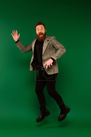 Excited bearded man in jacket with clover waving hand and jumping on green background 