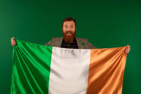 Happy bearded man holding Irish flag and looking at camera isolated on green 