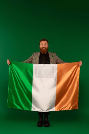 Longitud total del hombre barbudo positivo sosteniendo bandera irlandesa sobre fondo verde 