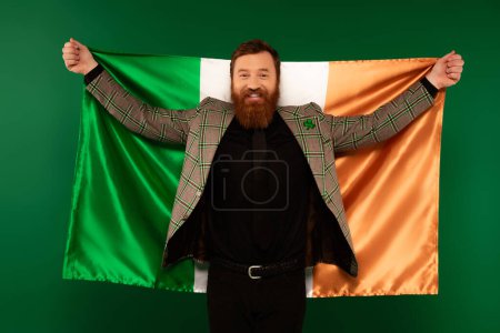 Smiling bearded man with clover on jacket holding Irish flag isolated on green 