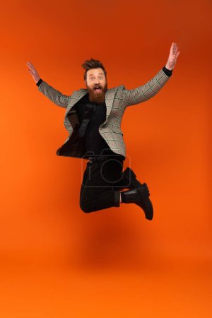 Hombre barbudo sorprendido en chaqueta a cuadros saltando sobre fondo rojo 