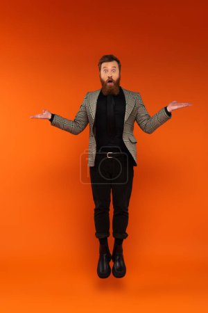 Shocked bearded man in blazer pointing with hands while jumping on red background 