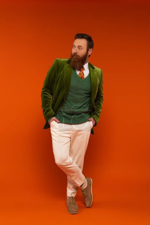 Trendy bearded man holding hands in pockets of pants on red background 