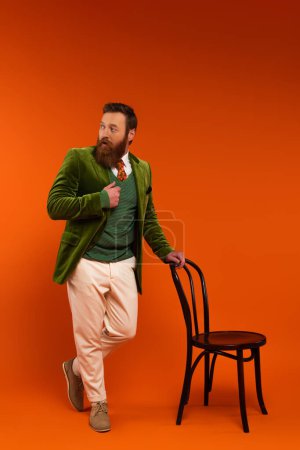 Longitud completa del hombre con estilo en la chaqueta de pie cerca de la silla sobre fondo rojo 