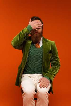Trendy bearded man in jacket covering eyes while sitting on chair on red background 