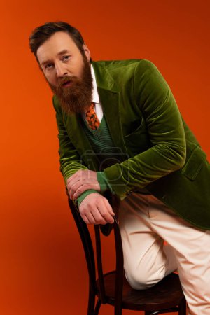 Modelo barbudo de moda posando en silla de madera sobre fondo rojo 