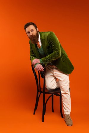 Full length of stylish bearded model posing near chair on red background 