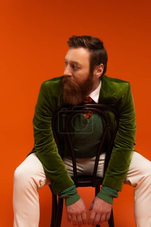 Trendy bearded man in green jacket sitting on chair on red background 