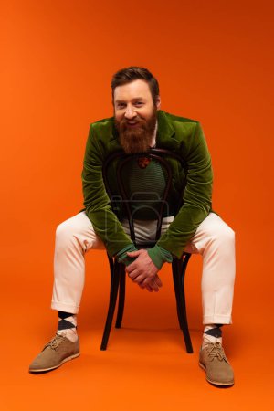 Full length of positive bearded man sitting on chair on red background 