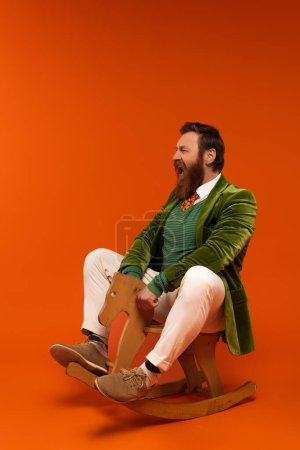 Excited bearded man in jacket sitting on rocking horse on red background 