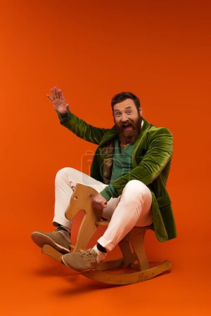 Excited bearded man in jacket waving hand while sitting on rocking horse on red background 