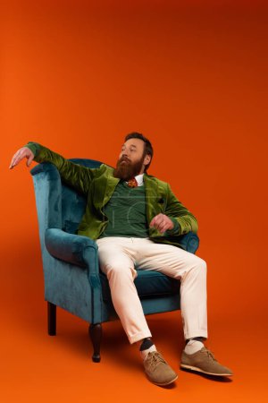 Stylish bearded man sitting on blue armchair on red background 