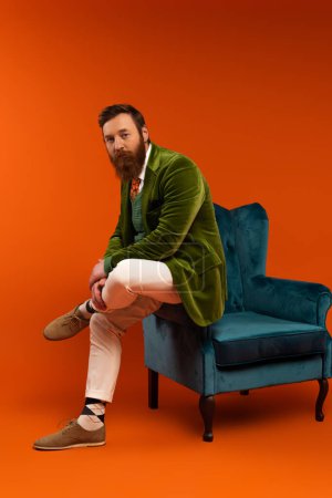 Trendy bearded model in stylish clothes sitting on blue armchair on red background 