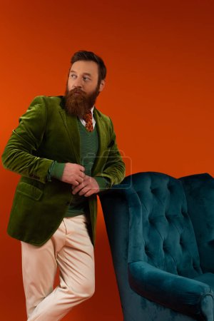 Hombre barbudo de moda mirando hacia otro lado cerca de sillón de terciopelo azul sobre fondo rojo 
