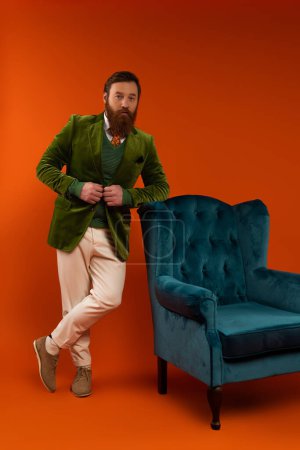 Trendy bearded model looking at camera near blue armchair on red background 
