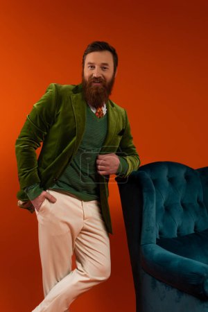 Stylish bearded man looking at camera while posing near armchair on red background 
