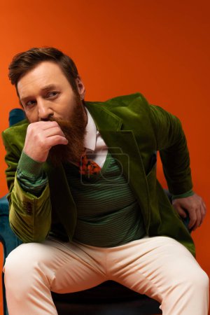 Fashionable bearded man in jacket sitting on armchair on red background 