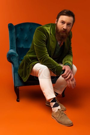 Full length of stylish bearded model looking at camera while sitting on armchair on red background 
