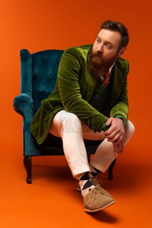 Trendy bearded man sitting on blue armchair on red background 