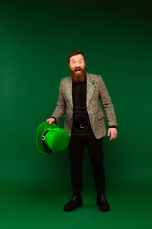Excited bearded man holding hat and looking at camera during saint patrick celebration on green background  tote bag #640348550