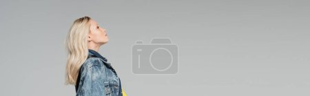 Photo for Side view of young blonde woman in stylish denim jacket looking away isolated on grey, banner - Royalty Free Image