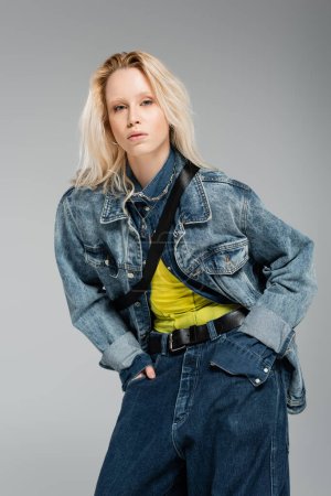 young blonde woman in stylish denim outfit looking at camera while posing with hands in pockets isolated on grey 