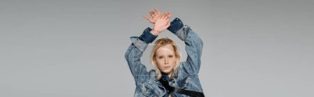 young blonde woman in stylish denim jacket looking at camera while posing with hands above head isolated on grey, banner 