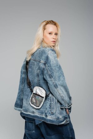 Foto de Young blonde model in stylish denim jacket with belt bag posing isolated on grey - Imagen libre de derechos