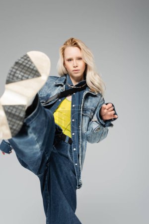 young blonde model in stylish denim outfit and trendy sneakers kick posing isolated on grey 