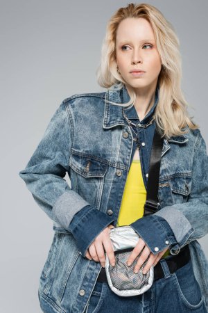 blonde albino model in stylish denim outfit with belt bag posing isolated on grey 