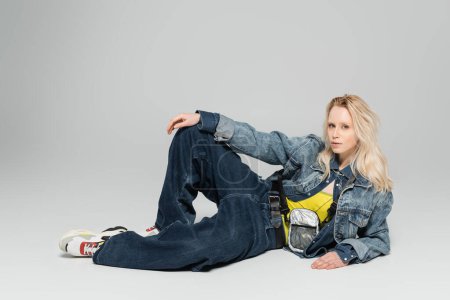 Photo for Full length of young blonde woman in blue denim outfit and trendy sneakers posing on grey background - Royalty Free Image